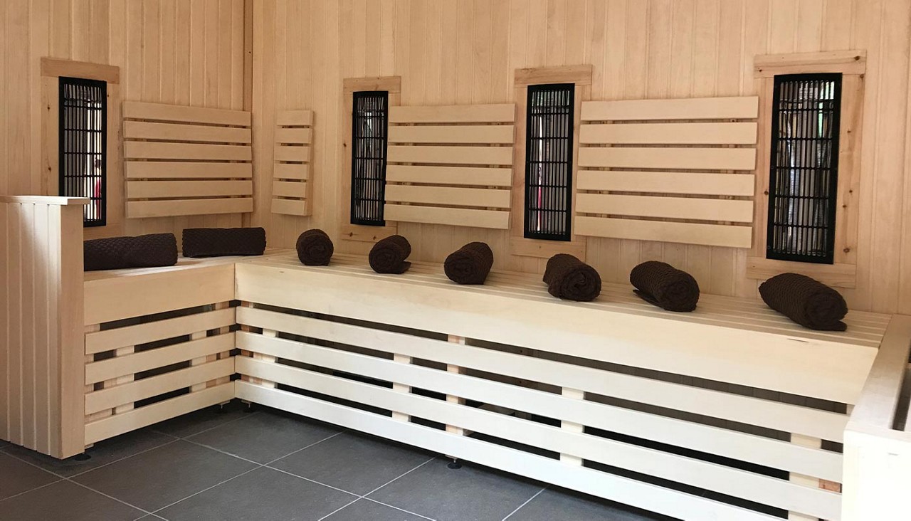 Wooden sauna benches display evenly spaced rolled brown towels, set against slatted backrests and vertical black heater panels, in a light-wood, corner infrared sauna with gray tiled floor.