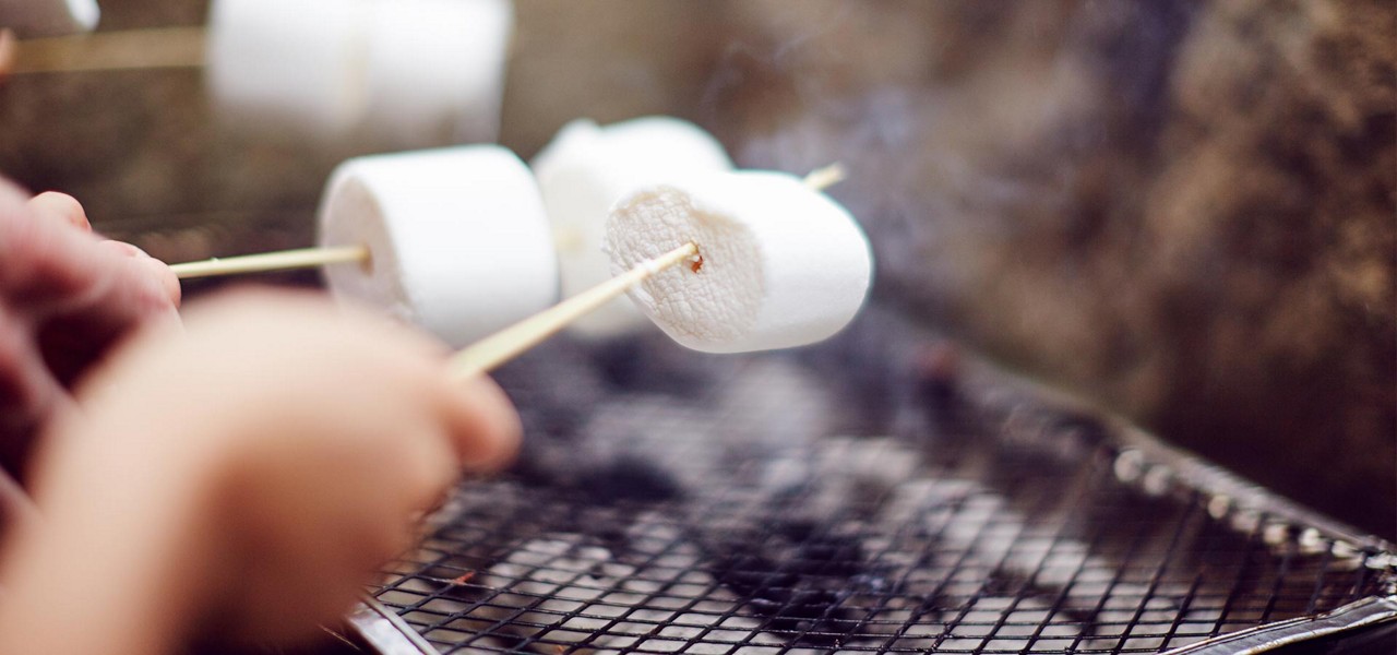 Marshmallows roast on wooden skewers, held by hands above a wire grill, faint smoke rising in an outdoor cooking setting.